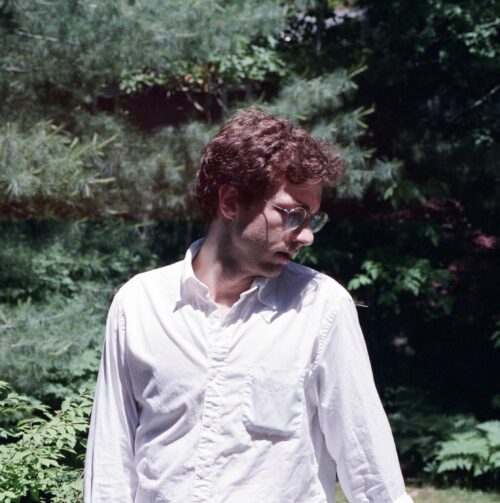 An image of Noah Rawlings, a person with short, red hair in a button-up shirt looking away from the camera